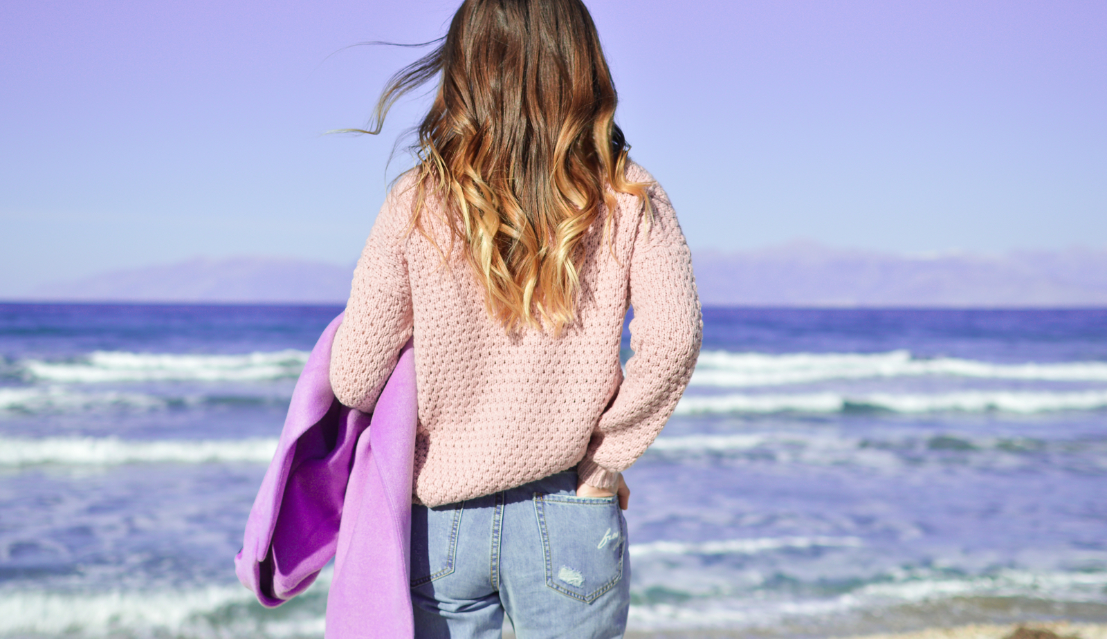 Woman with ombre hair wearing rose pink sweater holding lilac coloured sweater with one arm, other hand in pocket, looking out at the ocean